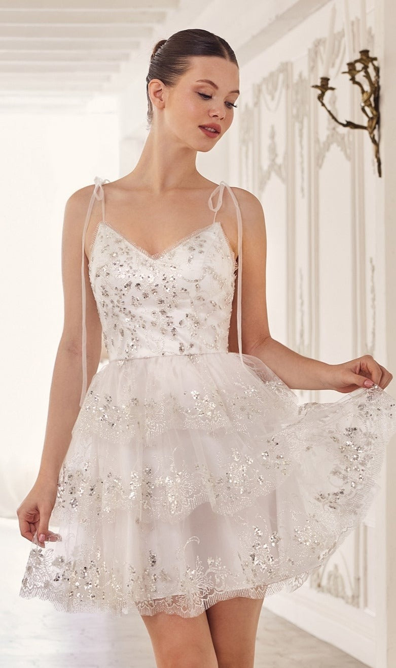 A woman wearing the Sequin-Print Short Ruffled Homecoming Dress KV1090 with floral embroidery and silver sequins stands indoors by a white ornate wall.